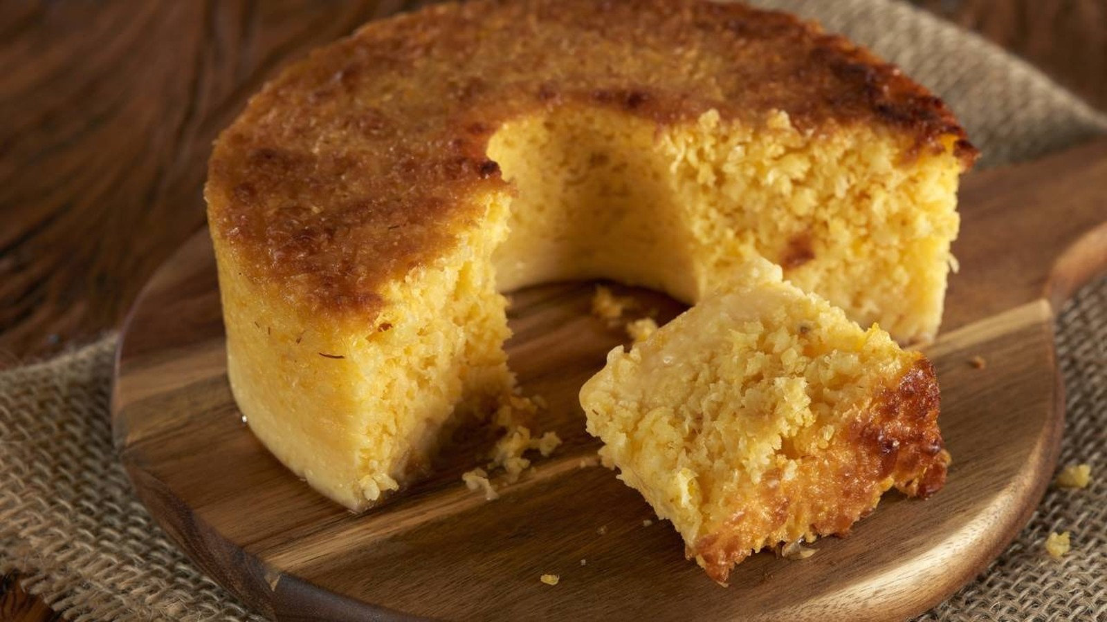 Bolo de fubá cremoso e dourado com uma fatia removida, sobre uma tábua de madeira