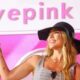 Mulher sorridente com chapéu preto e braço levantado sob o letreiro "wepink" rosa