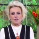 Ana Maria Braga com cabelo loiro, maquiagem escura e colar, em estúdio com plantas