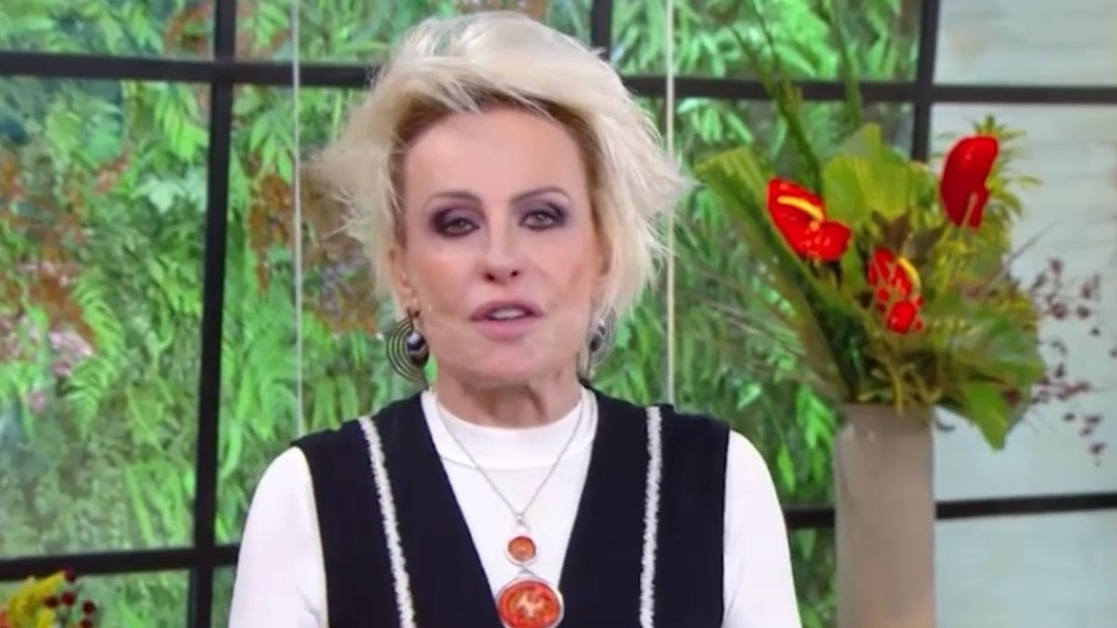 Ana Maria Braga com cabelo loiro, maquiagem escura e colar, em estúdio com plantas