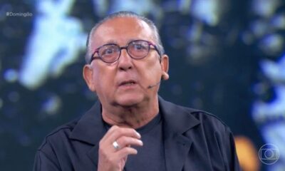 Galvão Bueno, de óculos, microfone e anel, fala em estúdio de TV. #Domingão