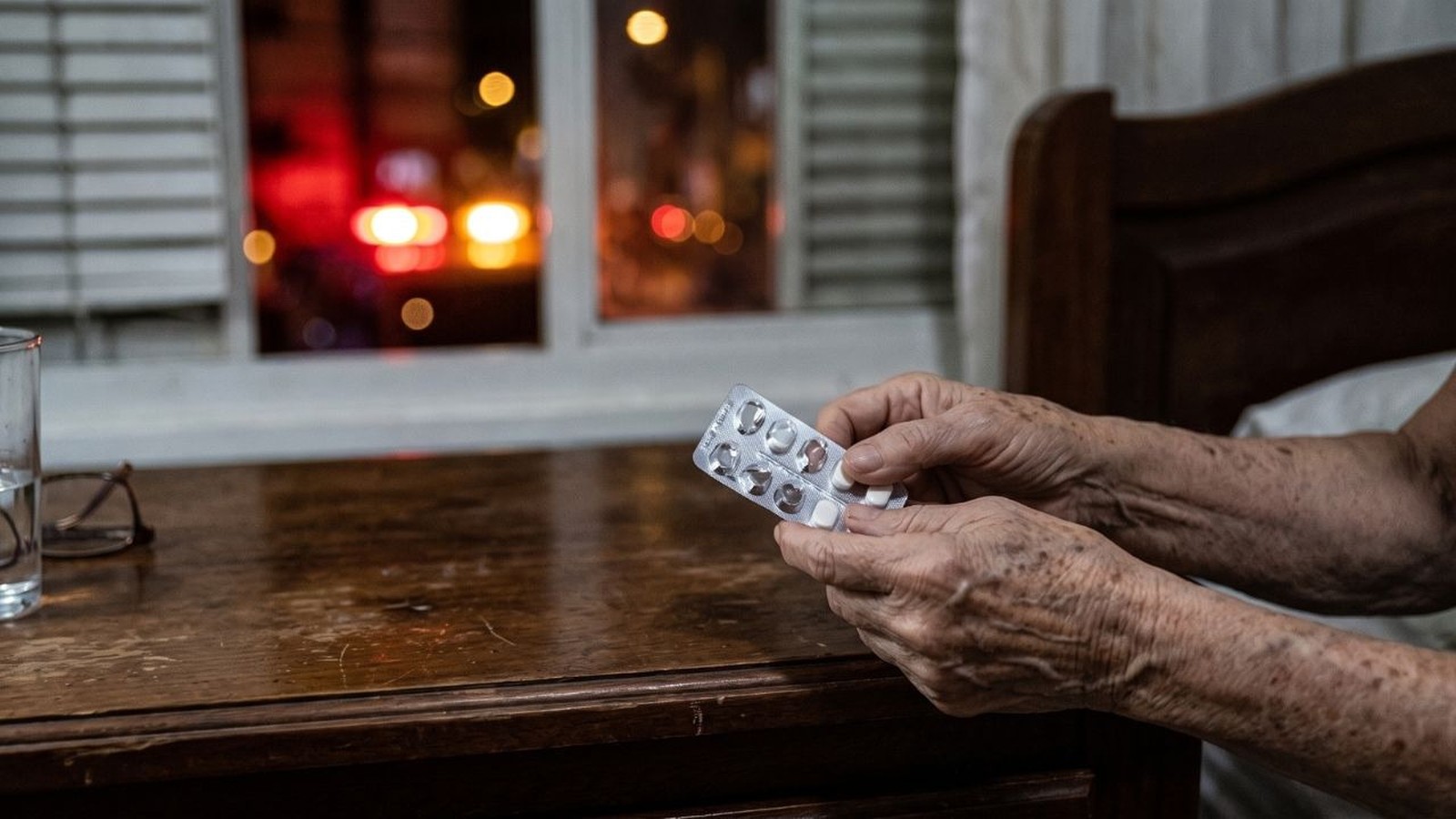 Mãos de idosa seguram cartela de remédios; copo d'água e janela com luzes noturnas