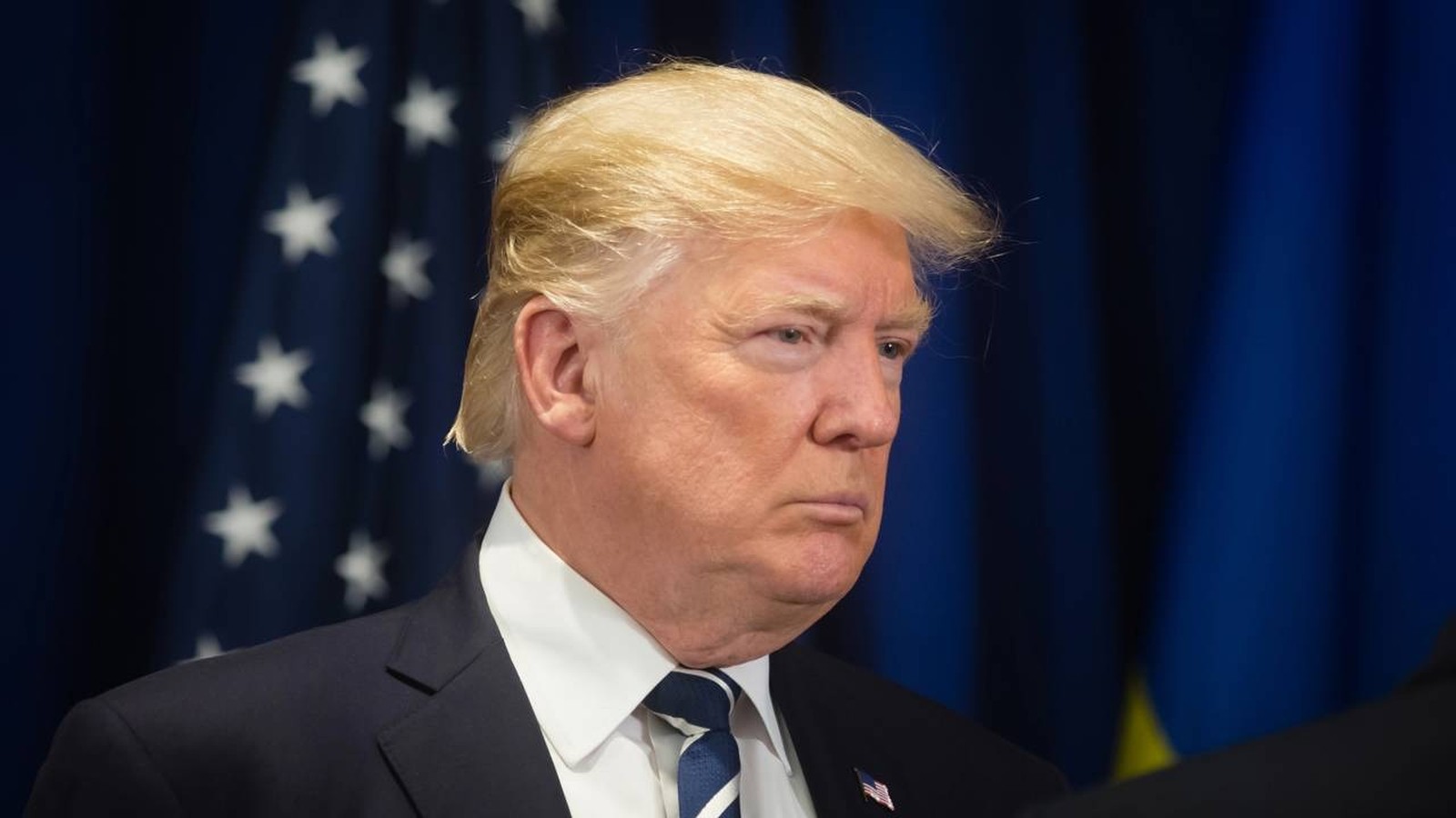 Close-up de Donald Trump com terno escuro e gravata em frente a bandeira dos EUA