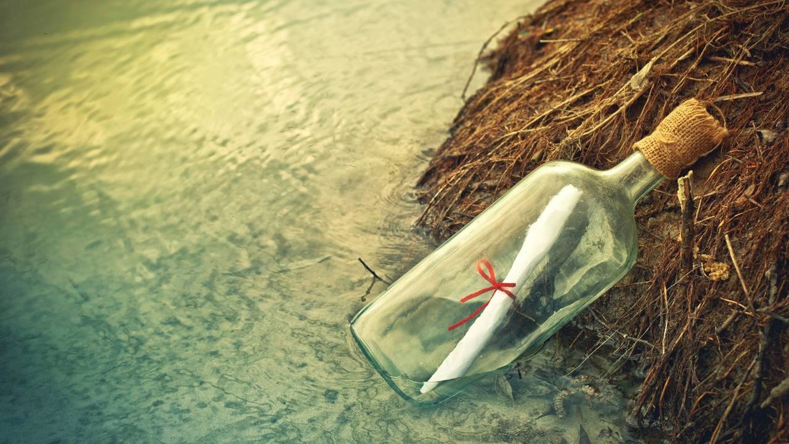 Garrafa de vidro com mensagem de papel enrolado e fita vermelha na água e areia