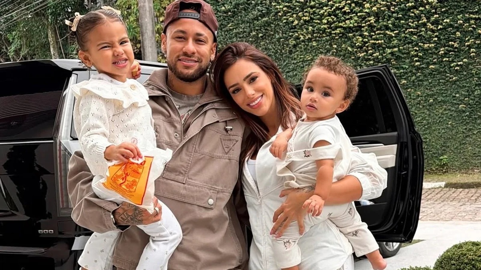 Neymar, Bruna Biancardi, Mavie e Mel posam juntos sorrindo, com carro e folhagem ao fundo