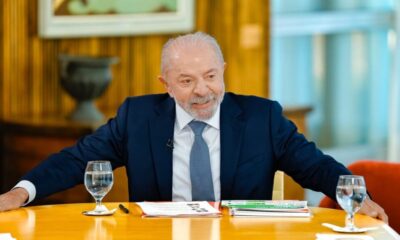 Luiz Inácio Lula da Silva sorrindo, vestindo terno azul, sentado à mesa com copos d'água
