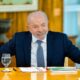 Luiz Inácio Lula da Silva sorrindo, vestindo terno azul, sentado à mesa com copos d'água