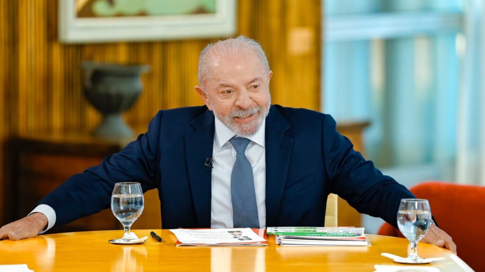 Luiz Inácio Lula da Silva sorrindo, vestindo terno azul, sentado à mesa com copos d'água