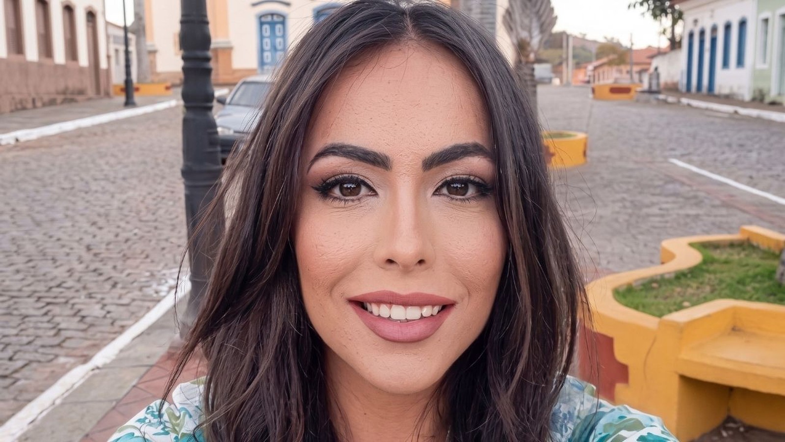 Selfie da mulher sorrindo com cabelo escuro e maquiagem, rua com paralelepípedos ao fundo