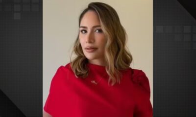 Mulher com cabelo castanho ondulado, blusa vermelha e colar dourado com um nome. Fundo claro