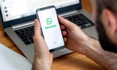 Mãos seguram celular exibindo logo e nome WhatsApp, com laptop desfocado ao fundo