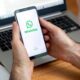 Mãos seguram celular exibindo logo e nome WhatsApp, com laptop desfocado ao fundo