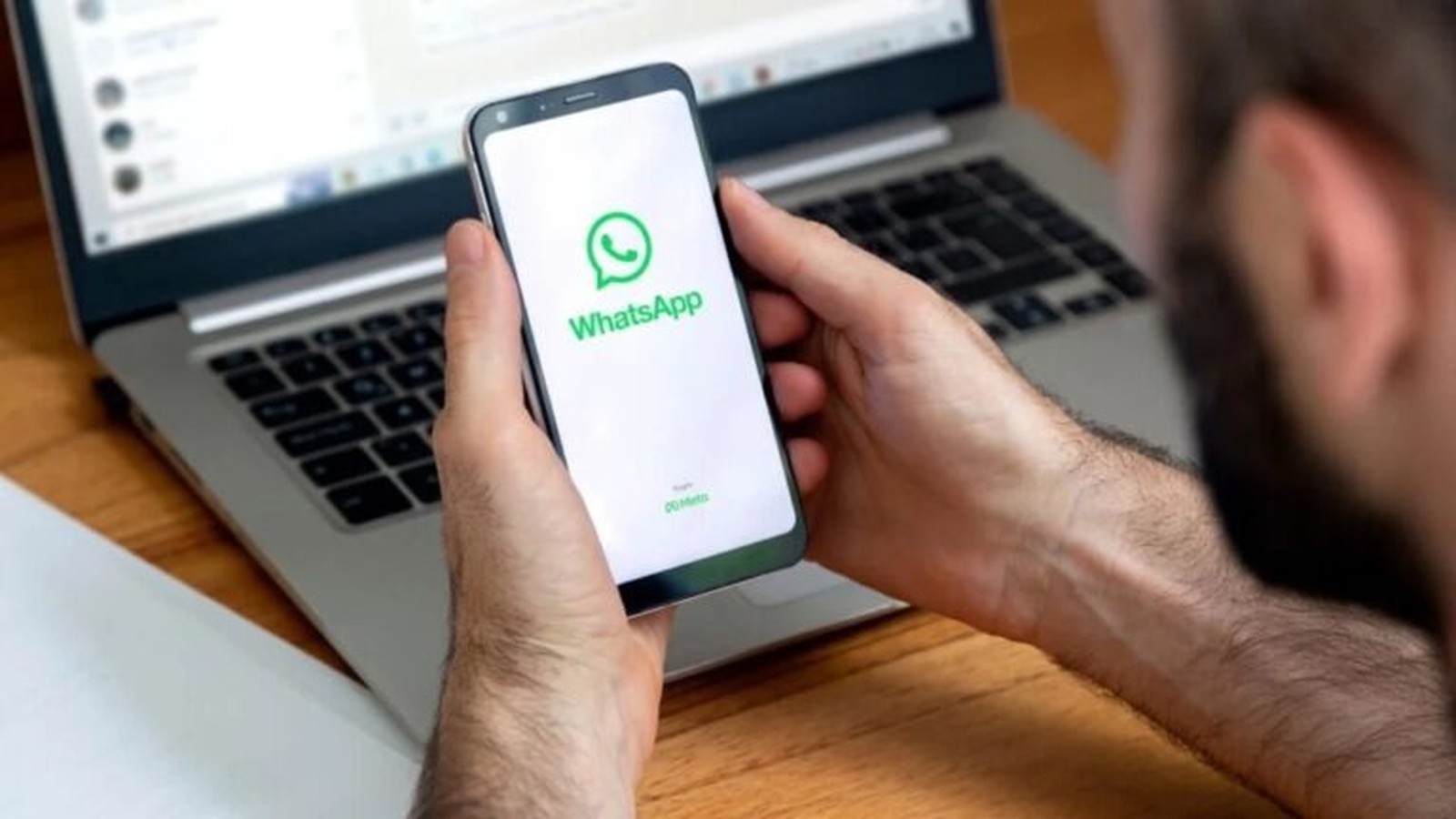 Mãos seguram celular exibindo logo e nome WhatsApp, com laptop desfocado ao fundo