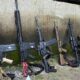 Cinco fuzis e uma mochila camuflada encostados em parede de concreto