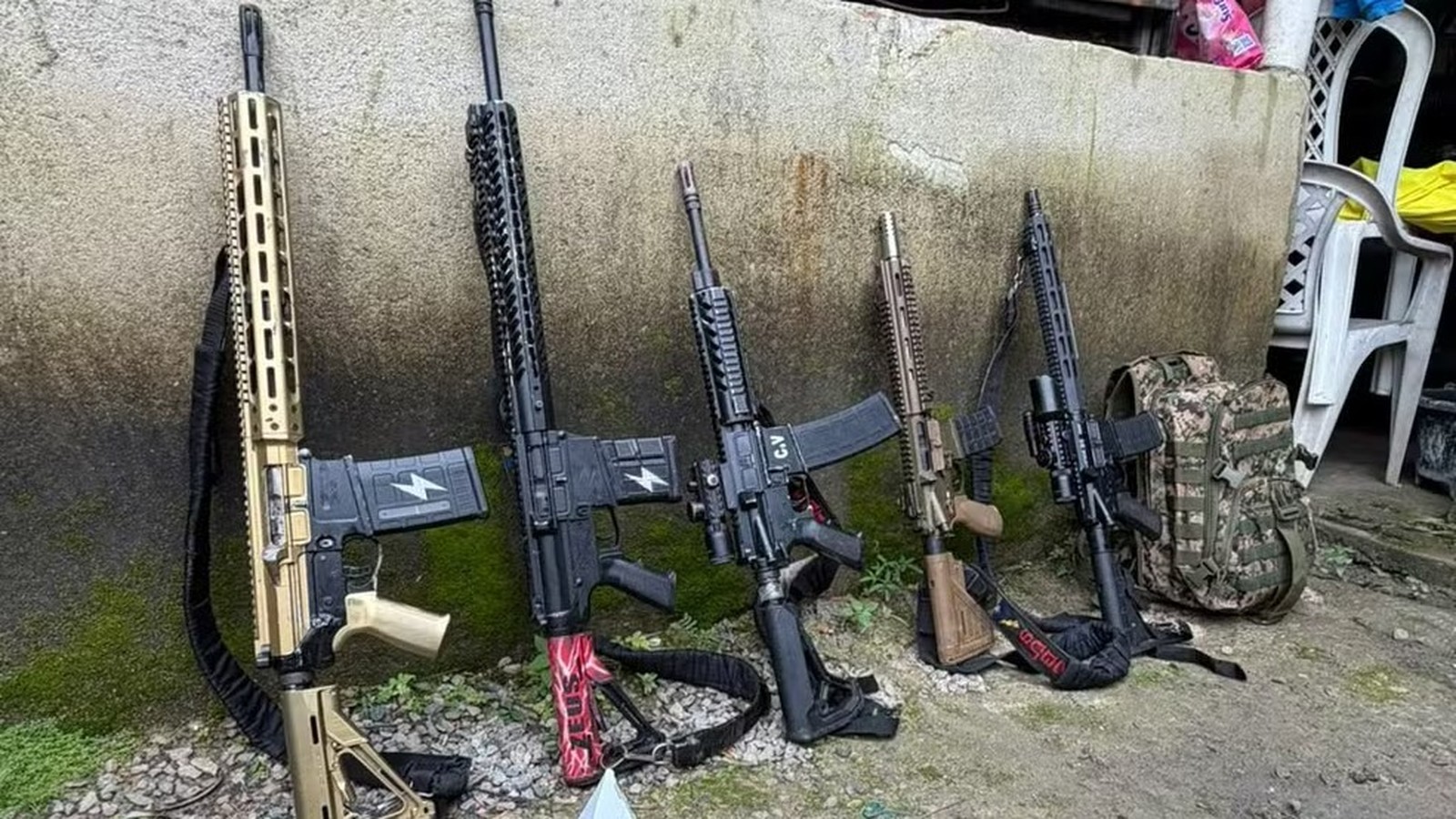 Cinco fuzis e uma mochila camuflada encostados em parede de concreto