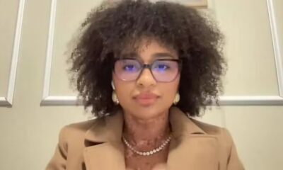Natália Deodato, mulher com óculos, cabelo crespo, blazer bege e colar no pescoço