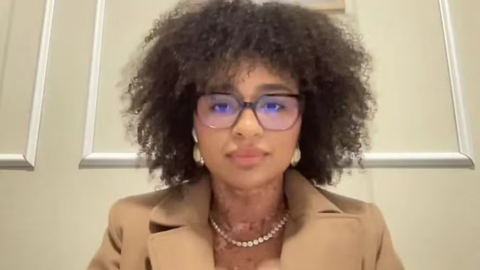 Natália Deodato, mulher com óculos, cabelo crespo, blazer bege e colar no pescoço