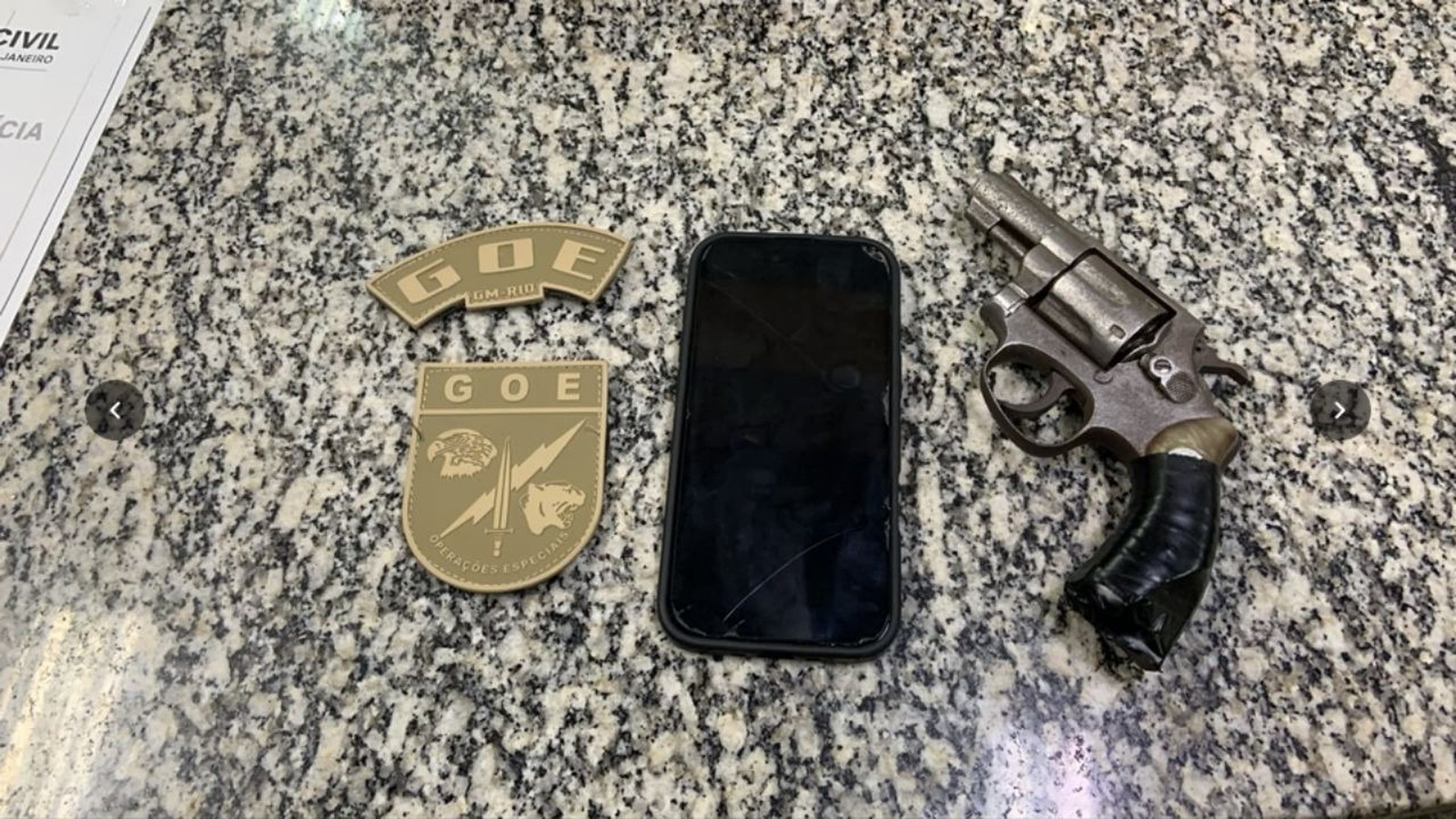 Dois distintivos do GOE, um celular preto com tela rachada e um revólver oxidado