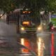 Ônibus e moto trafegam em rua alagada sob chuva forte, com ônibus espalhando água
