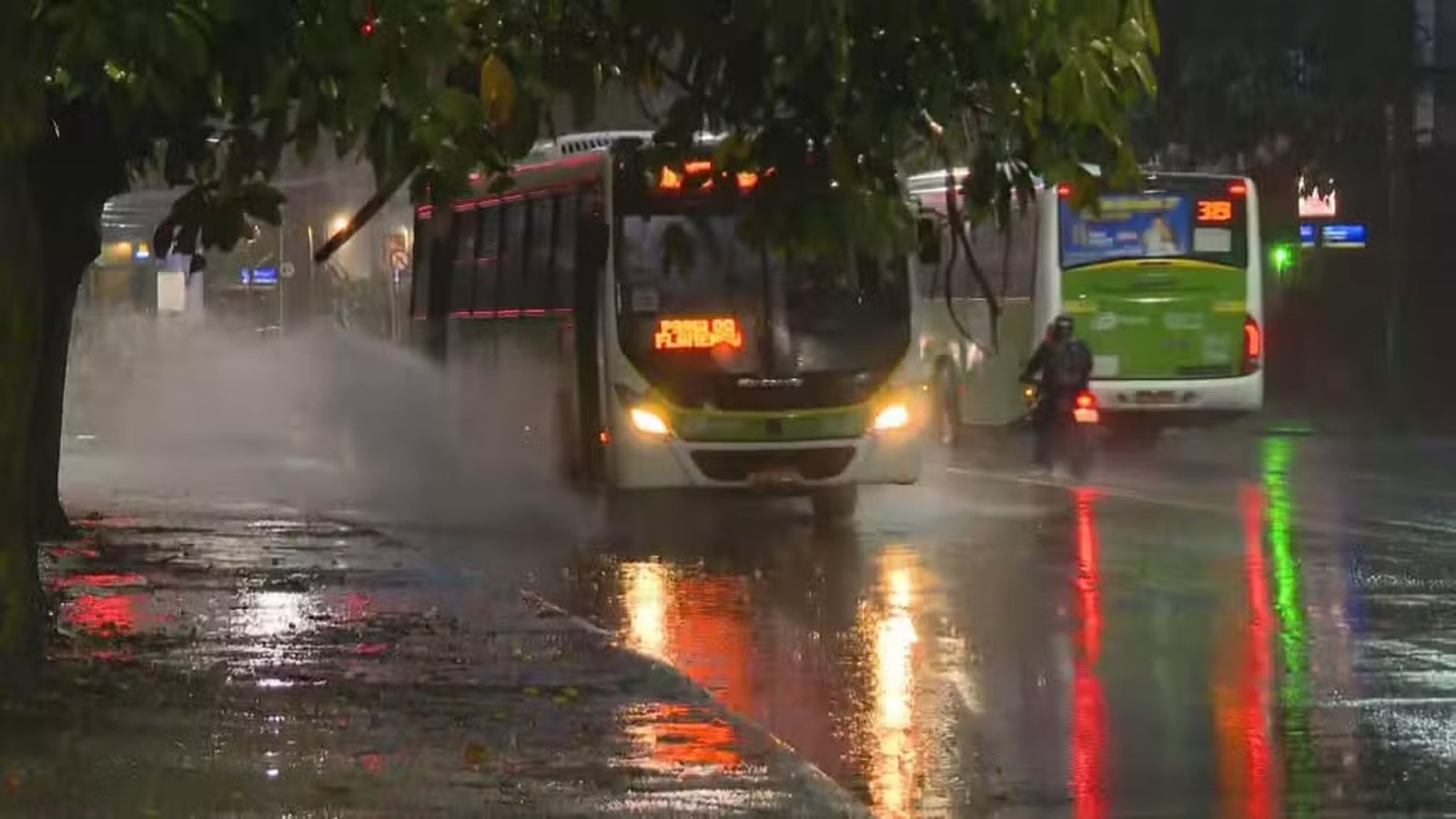 Ônibus e moto trafegam em rua alagada sob chuva forte, com ônibus espalhando água