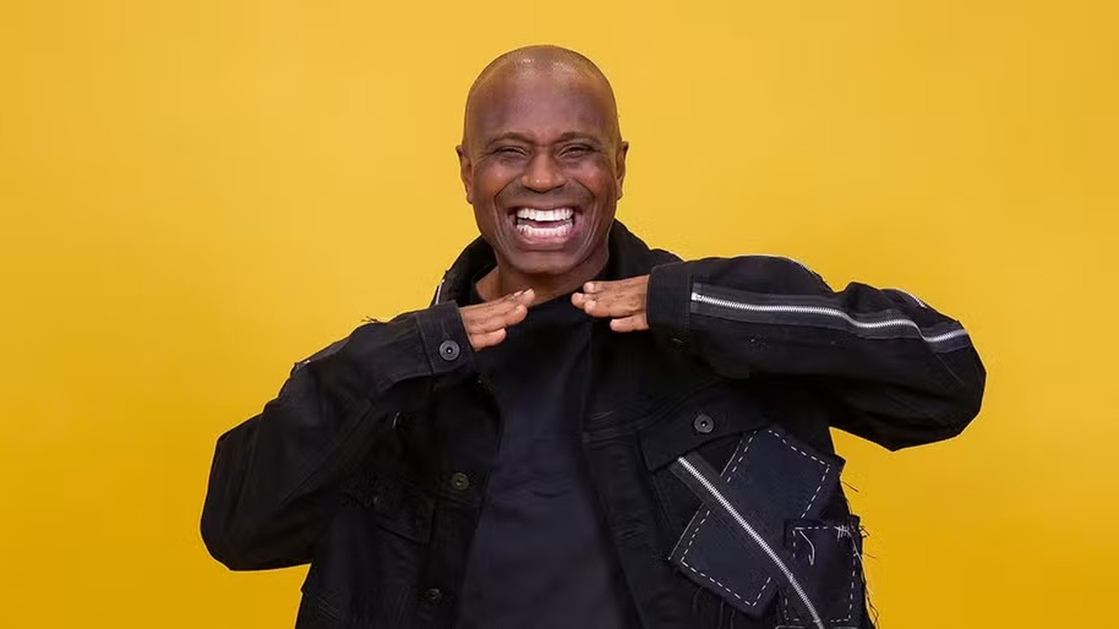 Homem negro careca sorrindo, mãos no peito, jaqueta preta em fundo amarelo