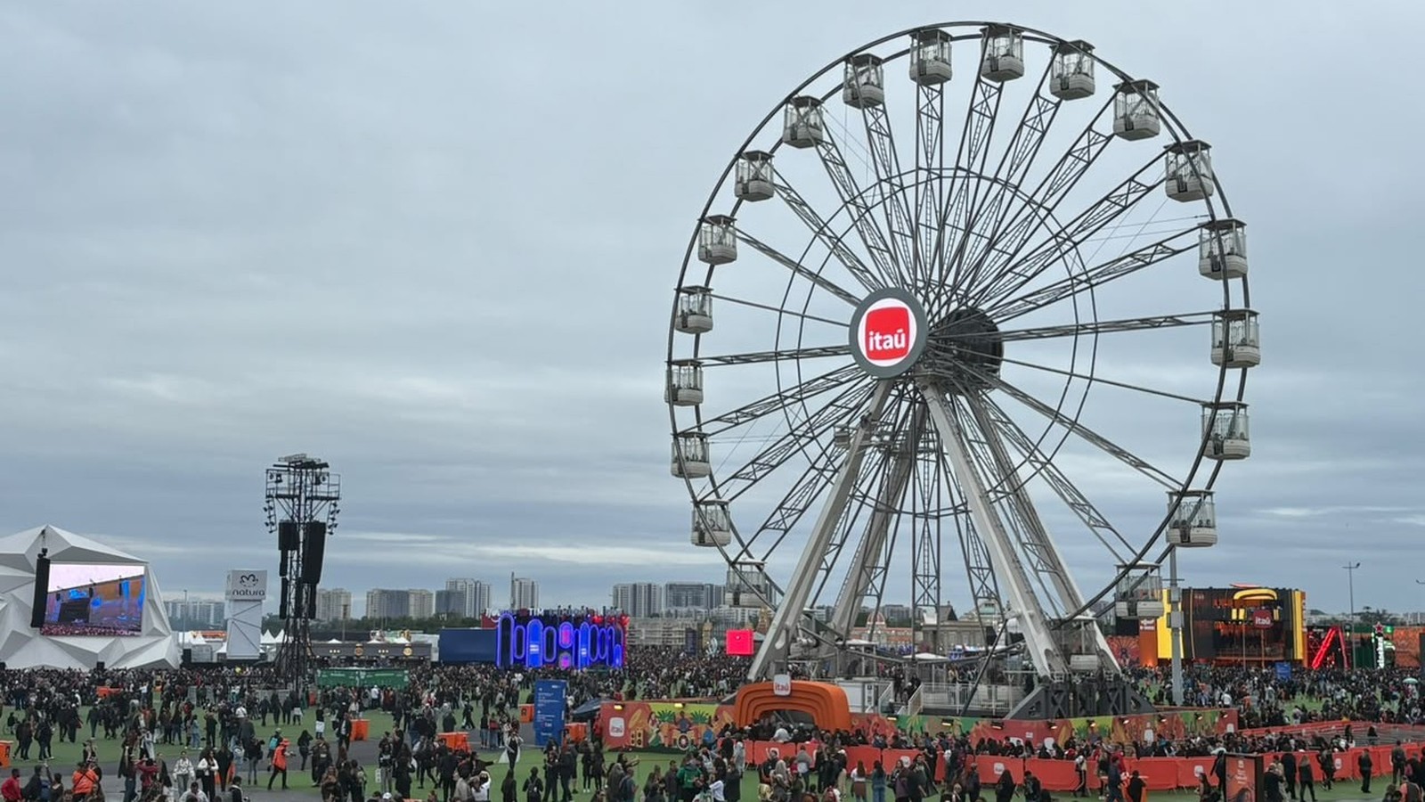 Uma roda gigante Itaú em um festival lotado com público e palcos sob céu nublado