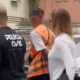 Agentes da Polícia Civil, um com camiseta ‘POLÍCIA CIVIL’, outro com colete, perto de viatura ‘26° DP’.