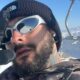Homem com óculos espelhados, gorro preto, barba e luvas em montanha nevada com teleférico