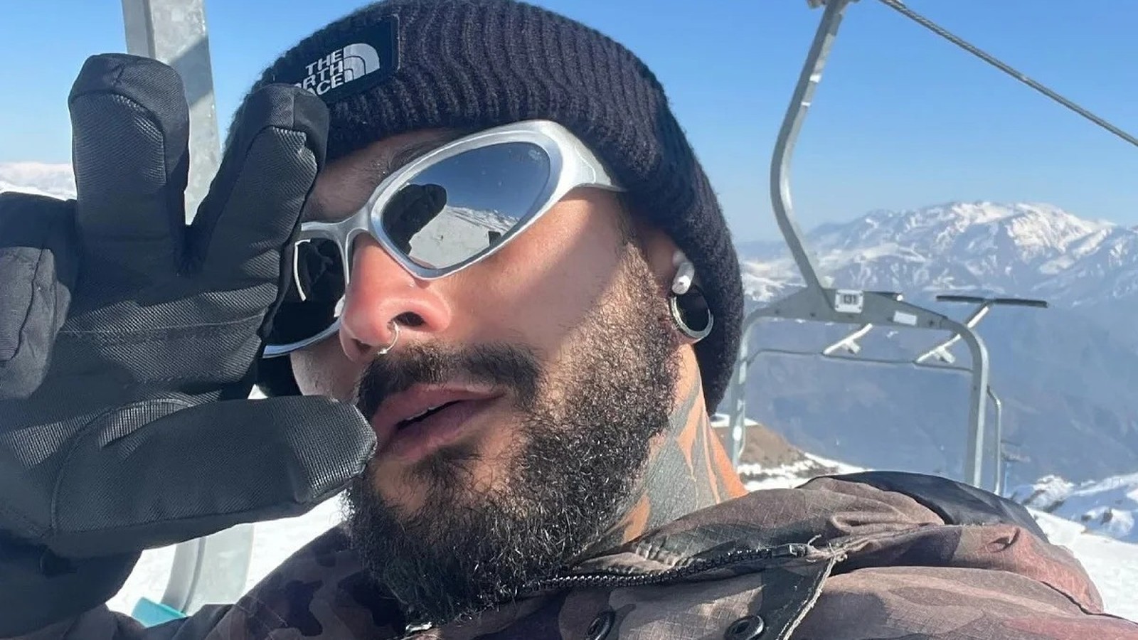 Homem com óculos espelhados, gorro preto, barba e luvas em montanha nevada com teleférico