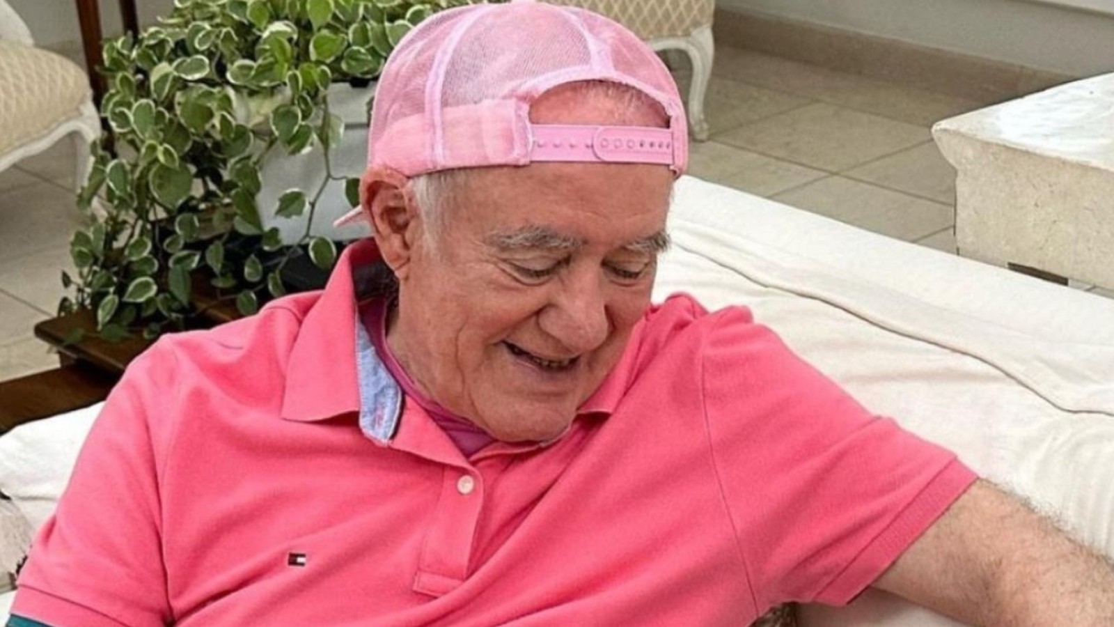 Renato Aragão de camisa polo rosa e boné rosa, sorrindo e olhando para baixo