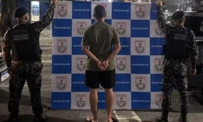 Homem algemado de costas flanqueado por dois policiais militares em frente a painel da Polícia Militar