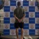 Homem algemado de costas flanqueado por dois policiais militares em frente a painel da Polícia Militar