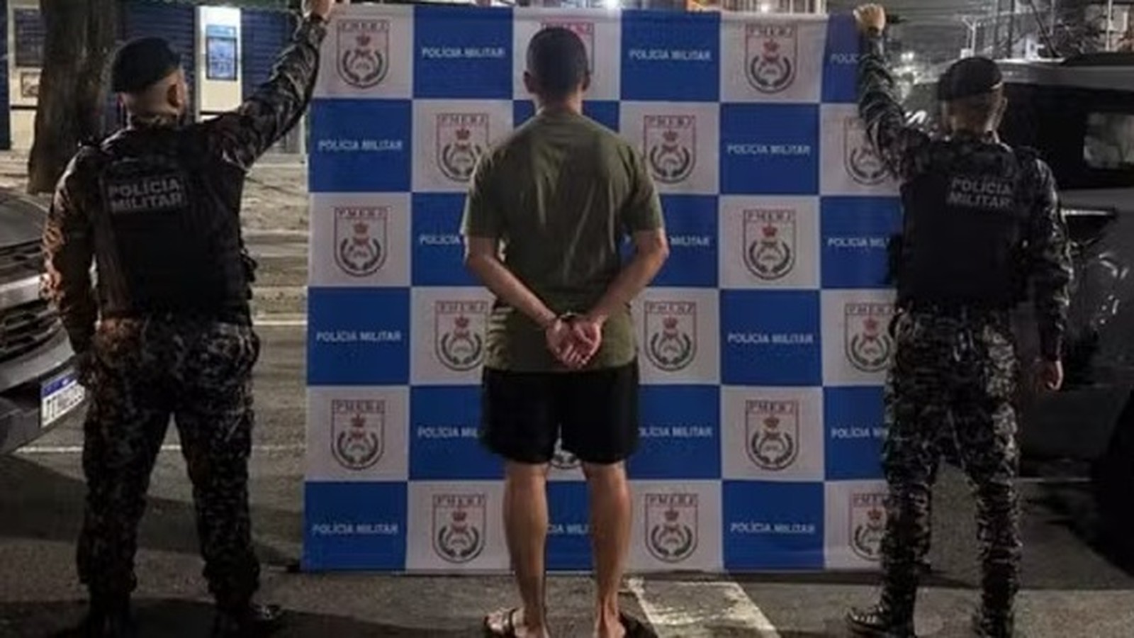 Homem algemado de costas flanqueado por dois policiais militares em frente a painel da Polícia Militar