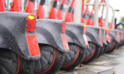 Patinetes elétricos cinza e vermelho enfileirados, com para-lamas molhados e luzes traseiras acesas