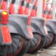 Patinetes elétricos cinza e vermelho enfileirados, com para-lamas molhados e luzes traseiras acesas