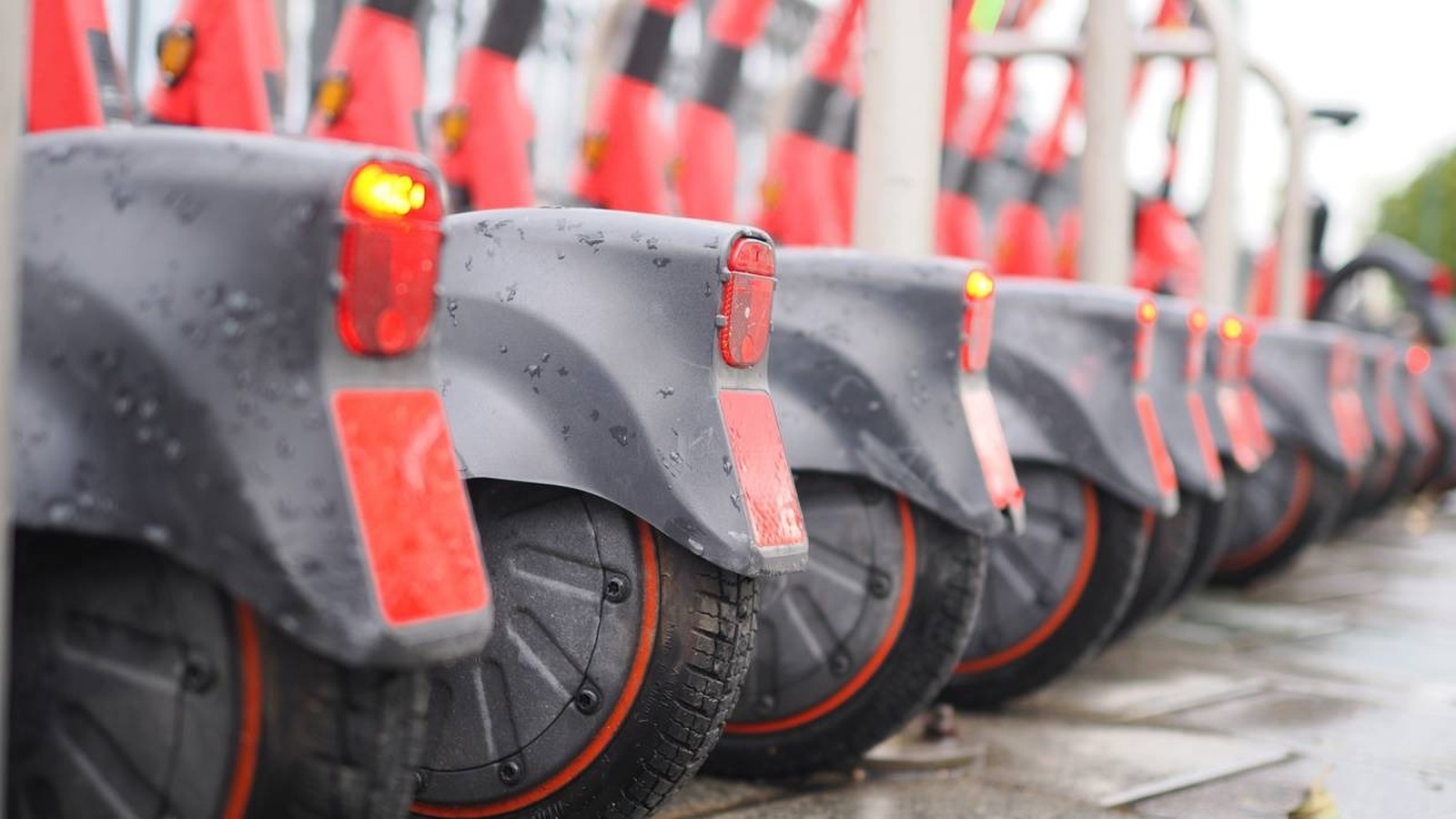 Patinetes elétricos cinza e vermelho enfileirados, com para-lamas molhados e luzes traseiras acesas
