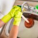 Mãos com luvas amarelas desentupindo pia de cozinha de inox com desentupidor e água