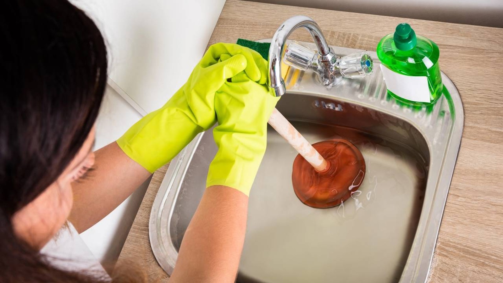 Mãos com luvas amarelas desentupindo pia de cozinha de inox com desentupidor e água