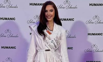Mulher sorrindo, vestindo blazer branco e faixa "AUSTRIA 2024", fundo com logotipos "HUMANIC" e "Silvia Schneider"