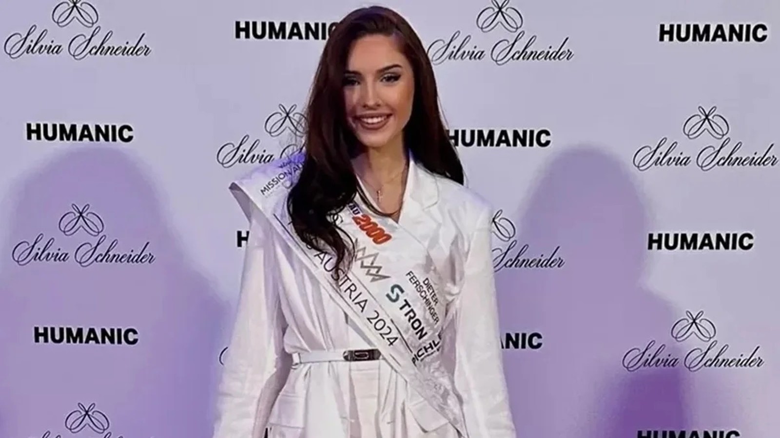Mulher sorrindo, vestindo blazer branco e faixa "AUSTRIA 2024", fundo com logotipos "HUMANIC" e "Silvia Schneider"