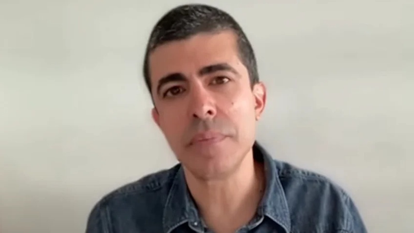 Homem com cabelo escuro e grisalho, camisa jeans azul, olhando para a câmera com expressão séria