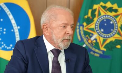 Luiz Inácio Lula da Silva, cabelo e barba brancos, terno azul e gravata, entre bandeiras do Brasil