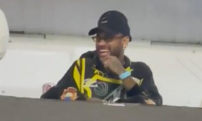 Neymar de boné, óculos e camisa preta e amarela rindo em um camarote