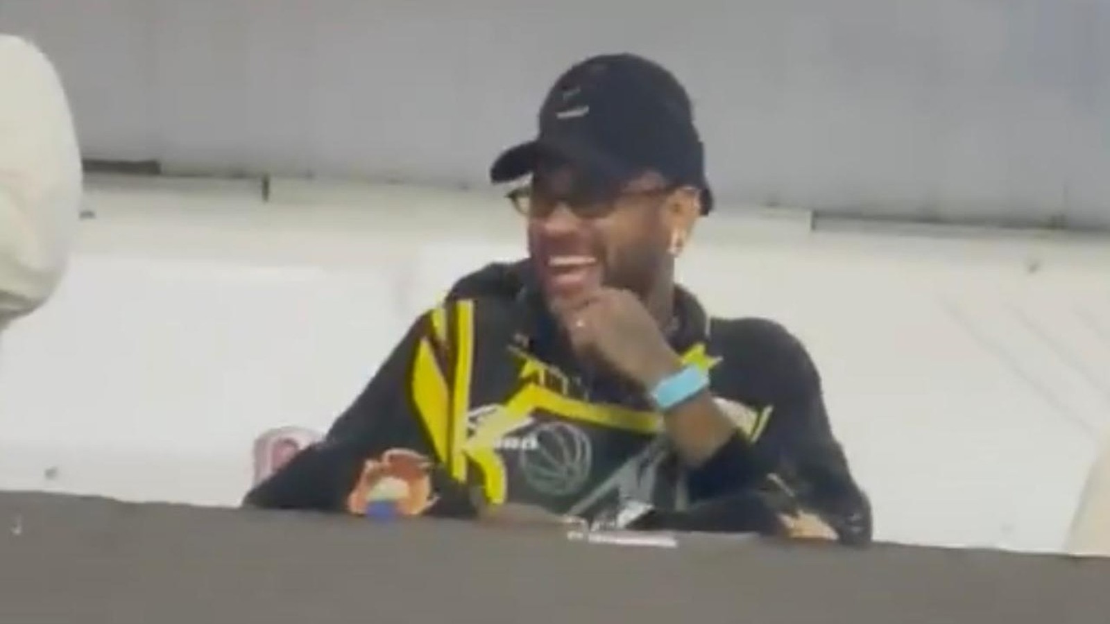Neymar de boné, óculos e camisa preta e amarela rindo em um camarote