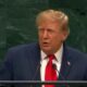 Donald Trump em um palanque, falando ao microfone, vestindo terno azul e gravata vermelha