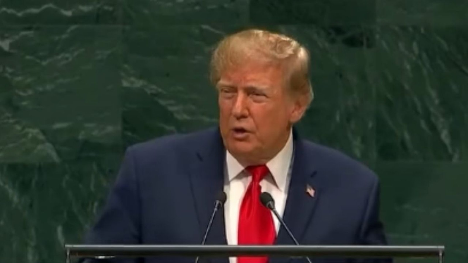 Donald Trump em um palanque, falando ao microfone, vestindo terno azul e gravata vermelha