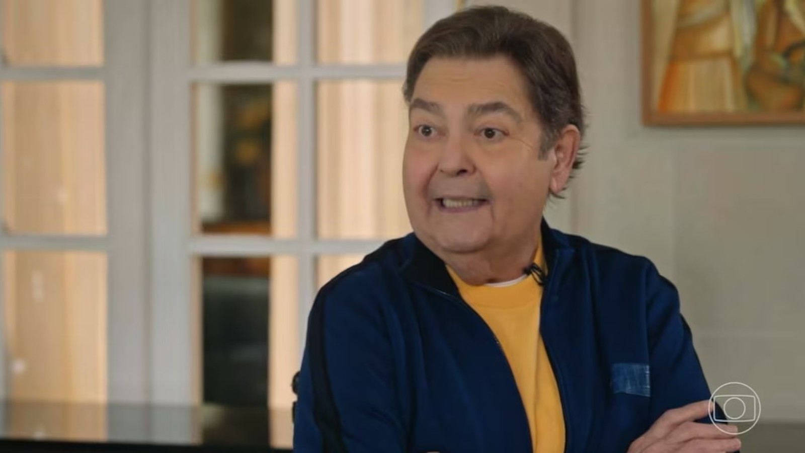 Fausto Silva em entrevista, jaqueta azul e camiseta amarela, com logo da Globo
