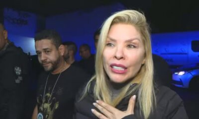 Mulher loira falando com mão no peito. Homem barbado, policiais e viatura policial ao fundo