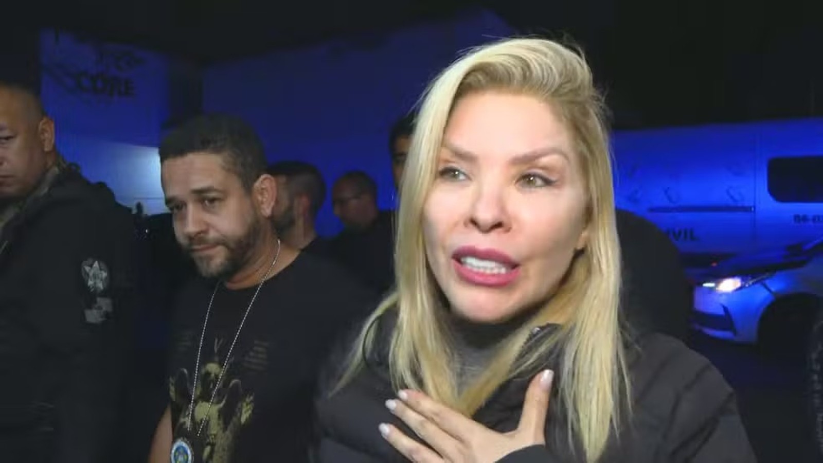 Mulher loira falando com mão no peito. Homem barbado, policiais e viatura policial ao fundo