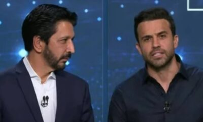 Dois homens em estúdio de TV, um de terno e outro de camisa escura, ambos com microfones